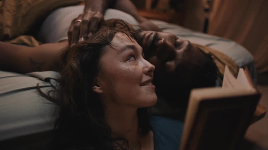 Woman reclined on the bed reads a book to her couple in the bedroom