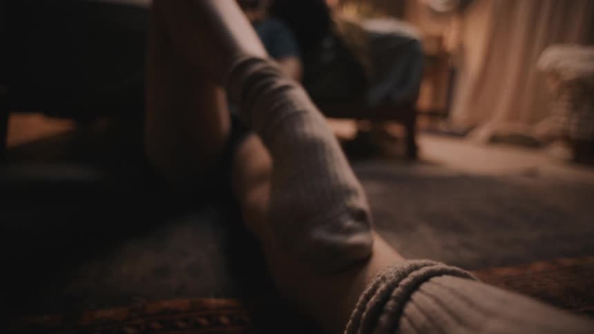 A woman sits at home and scratches her leg with her foot