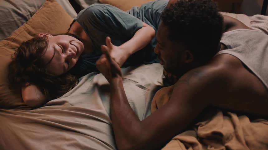 A man and woman are laughing while laying in bed playing thumb war
