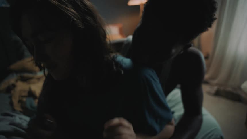 A man and woman sit on a bed and he holds her as she puts her arms up to push him away
