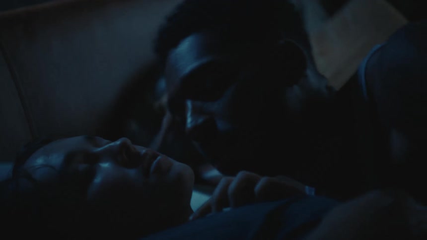 A man leans over a woman sleeping in bed and she puts her finger to her mouth to tell him to be quiet and pushes him away