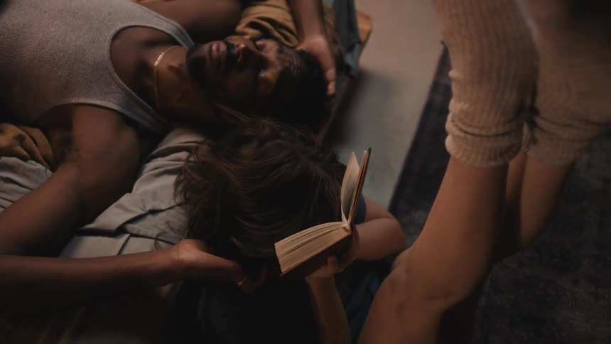 Woman reclined on the bed reads a book to her couple in the bedroom