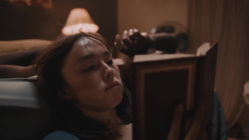 A woman is sitting on her bedroom floor leaning against the bed as she reads a book