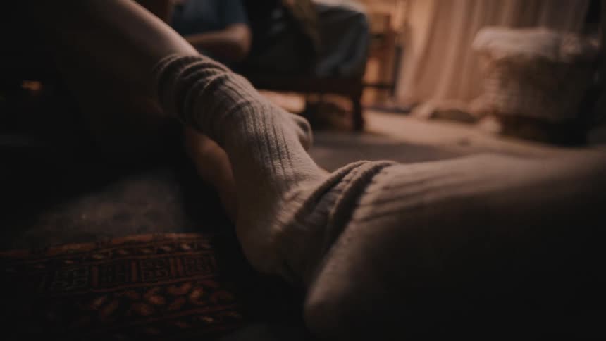 A person wearing long socks sitting on the floor rubbing legs with a foot
