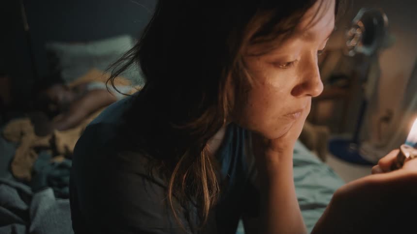 A teenage girl sits on a bed with a man sleeping on it and lights a lighter as she looks at it