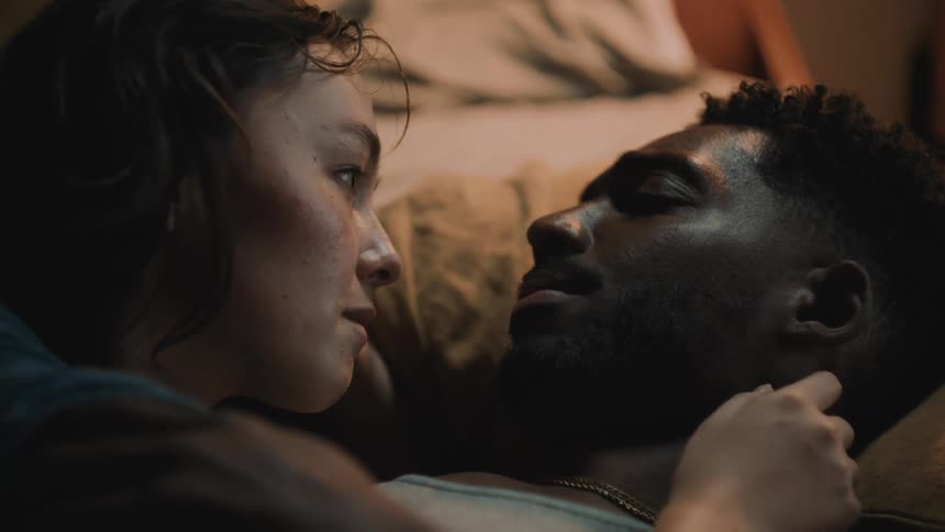 A man and woman lie in bed and cuddle and talk as she strokes his face