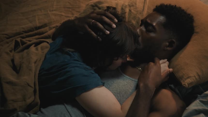 A man and woman lie in bed and hold hands as he strokes her hair and she kisses his chest