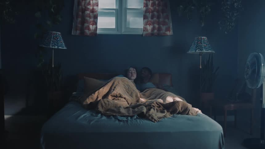 A woman gets into a bed and is laying next to a man under a blanket