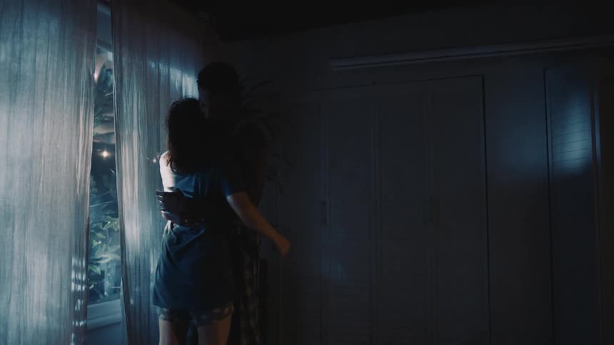 A man and woman in their sleeping clothing slowly dance before a window in a dark room at night
