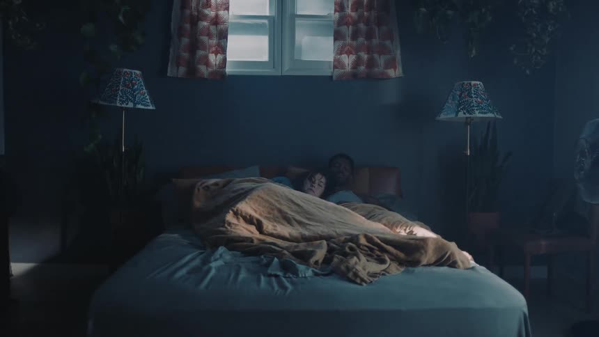 A woman climbs into bed under a blanket next to a man who is sleeping