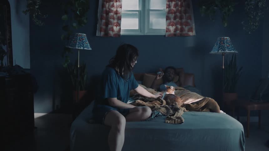 Woman with wavy hair black t shirt and shorts pets brown cat on bed lying next to man in bedroom