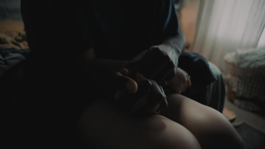 A person reaches around another persons waist and caresses their hands while sitting on a bed