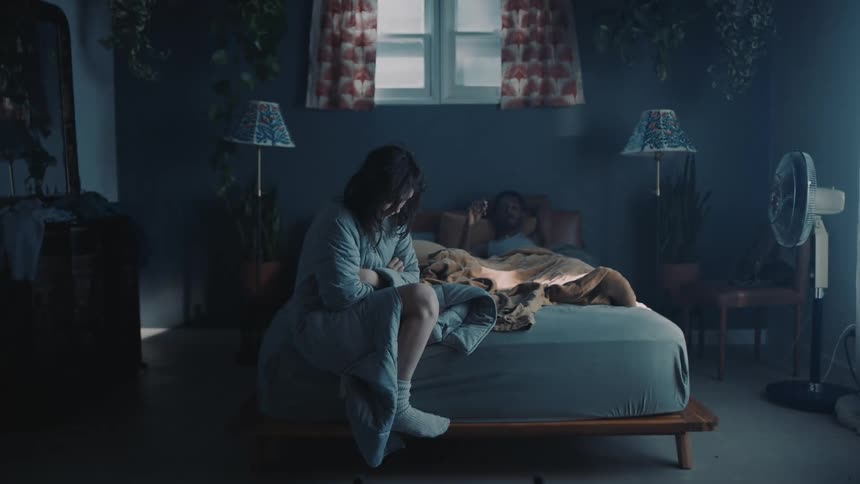 Woman sits downcast at foot of bed as man lies on it