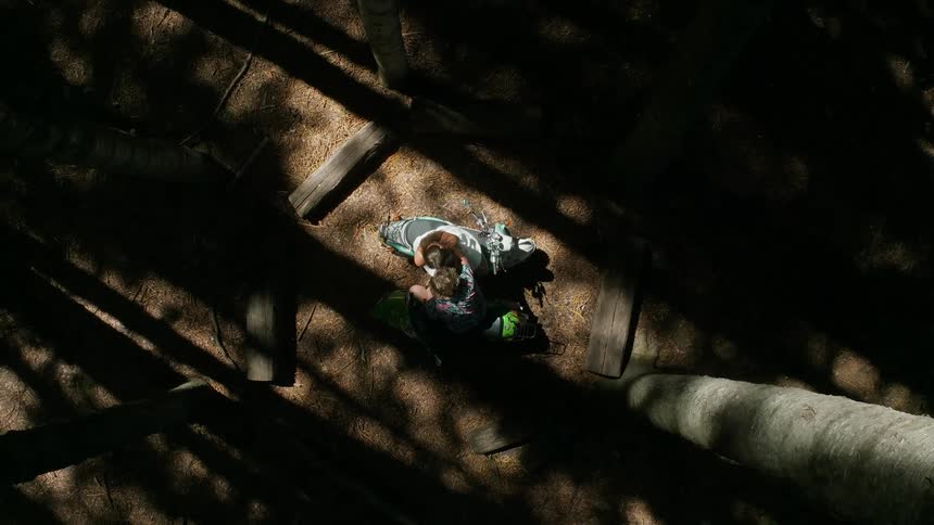 Two people sit on mopeds in a forest and lean towards each other to embrace