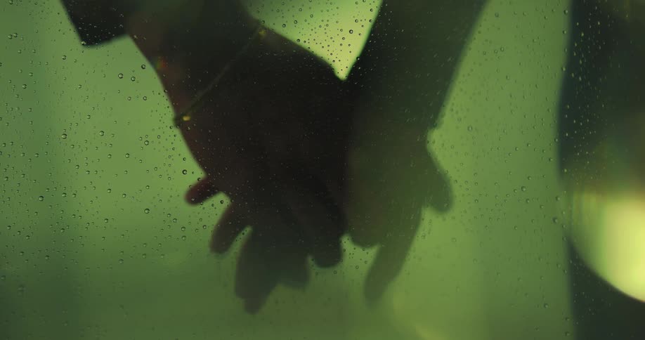 Close up of the silhouette of two people holding hands in front of a green glass