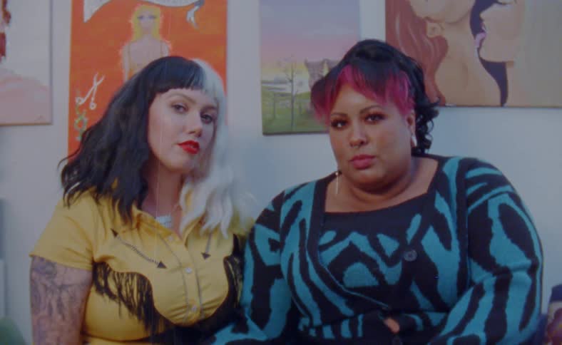 Two women with colorful hair sit together on a room looking at the camera