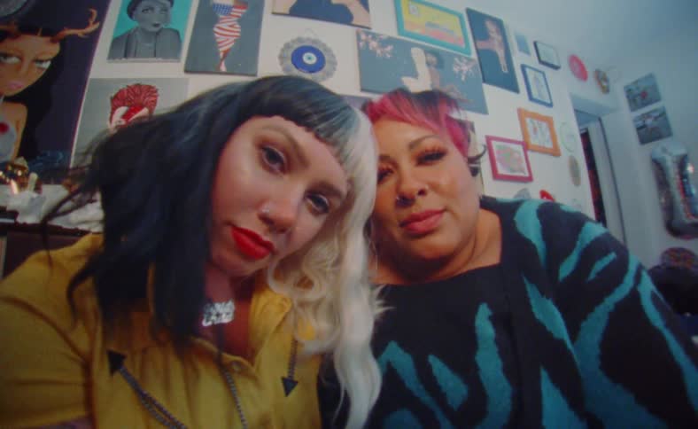 Two women with colorful hair sit togheter on a roomlooking at the camera