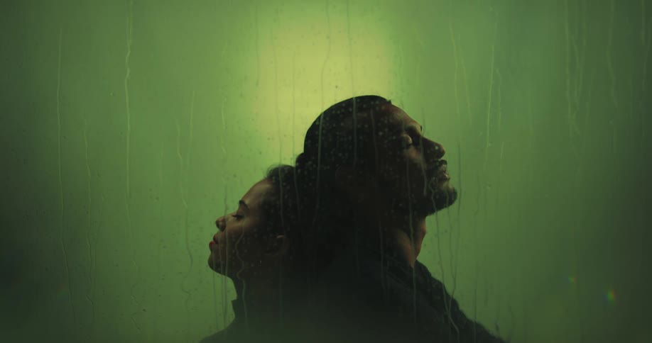 A couple sitting with their backs against each other behind a glass with water streaming down