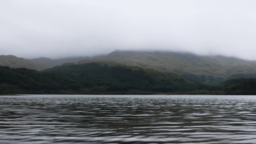 Light reflects on the water as it ripples and moves with mountains in the distance under a hazy sky