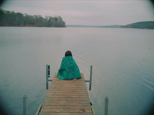 A person wrapped in a blanket on a lake dock at sunset