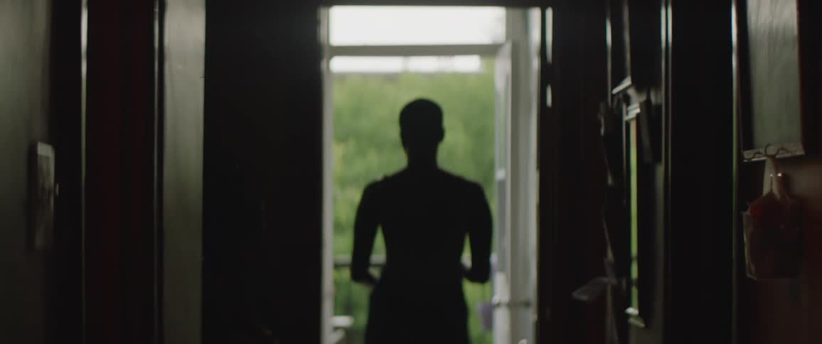 A woman walks down a corridor to exit a door onto a balcony before opening an umbrella