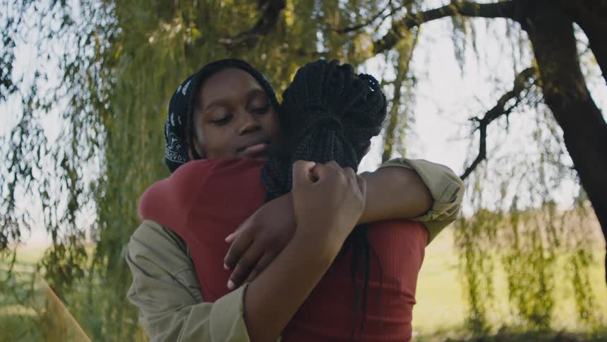 Two women hug one another under a large tree in a field