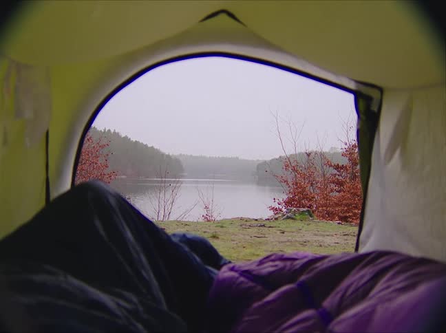 Two people are under blankets in a tent as they camp along the shore of a lake with a forest on the other side and mountains in the distance