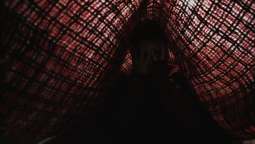 A child sitting under a blanket hides their face behind their hands