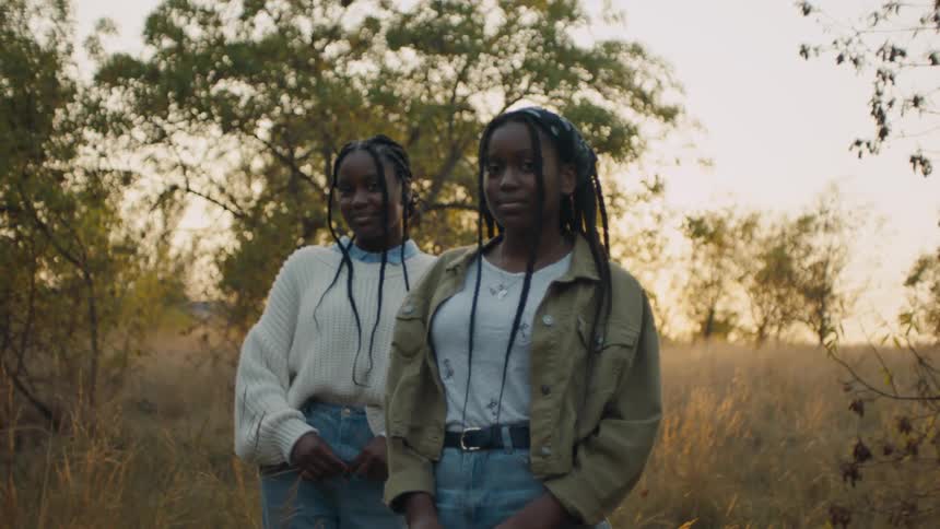 Two girls are standing outside in tall grass and weeds with trees