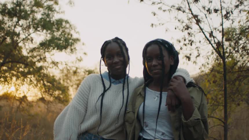Women with braids white knit sweater and white shirt and brown jacket smiles at camera at sunset in the field