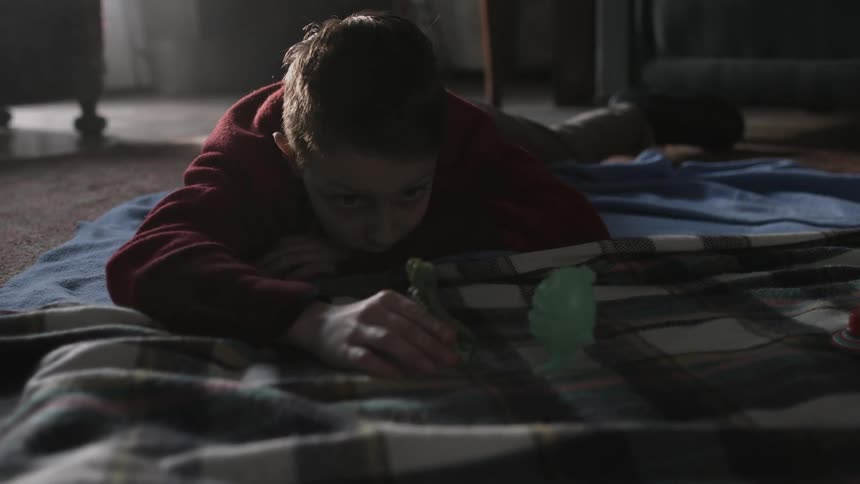 A bored looking boy lies on the floor at home and plays with a toy soldier