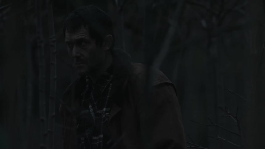 A man looks around at his surroundings with suspicion while standing in a woodland at night