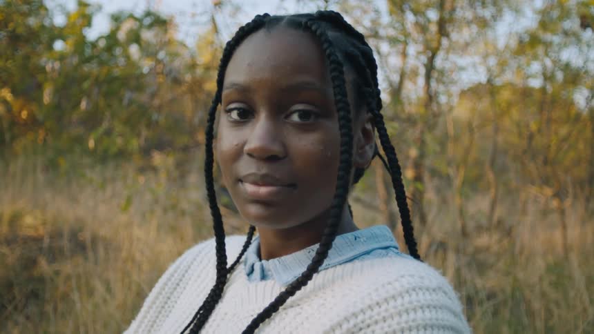 Woman with braids and white knit sweater looks at camera in the field