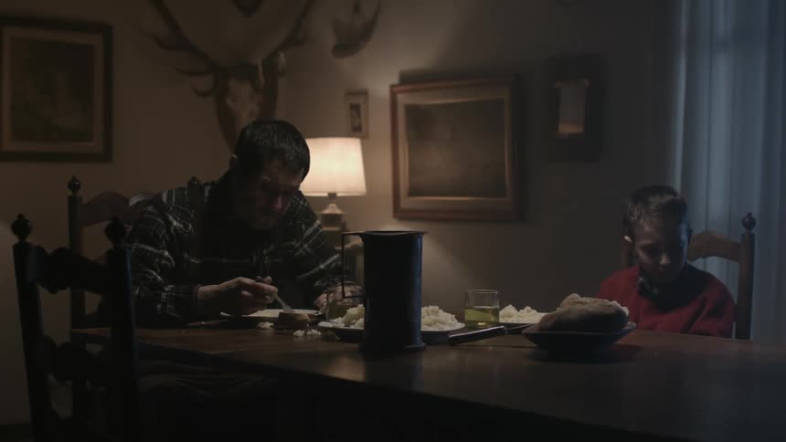 A sad looking boy sits at a table looking away from his food as his father eats and shakes his head at the boy