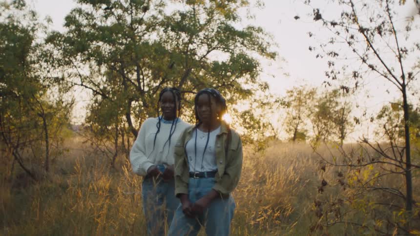 Two girls are standing outside in tall grass and weeds with trees in the distance