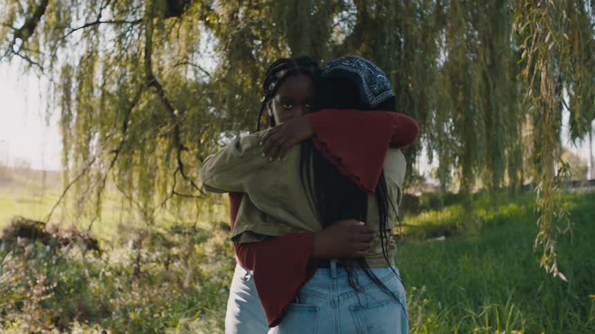 Two women stand hugging under the branches of a weeping willow tree