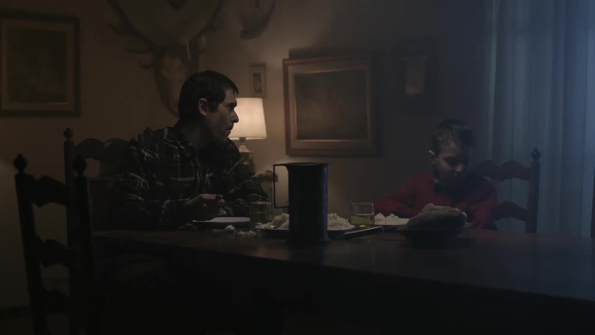 A sad looking boy sits at the dinner table and looks away from his plate while his dad eats and headlights shine in the window and they both get up