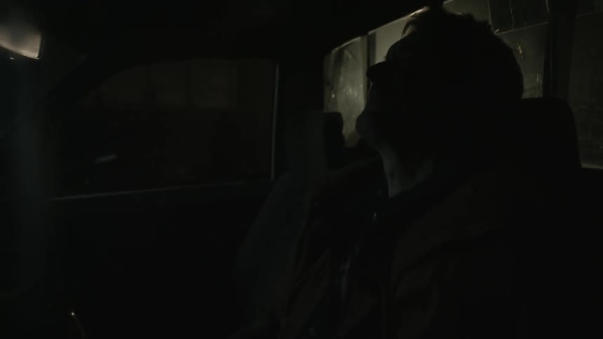 A man is sitting near windows in a dark room and he throws his head back as he speaks to someone