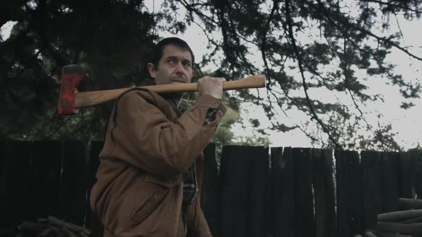 Nervous man holding an ax looking around next to a pile of logs in the backyard