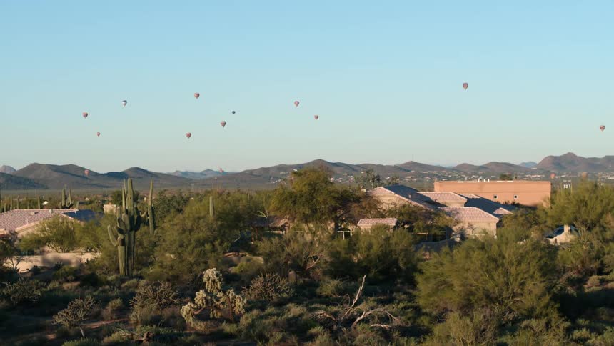 Hot air balloons float in a clear sky over a desert community with mountains in the distance