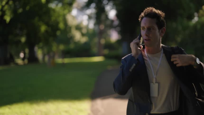 A man walks through a park on a phone and checks his watch