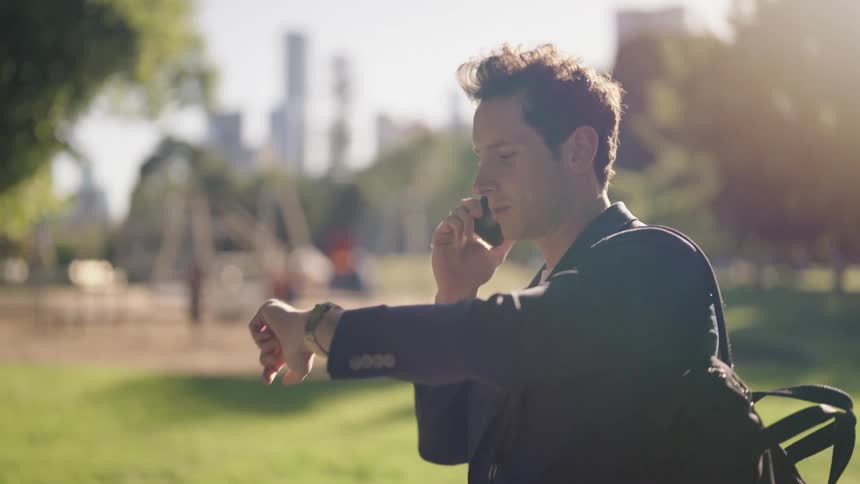A man is walking through a park holding the strap of his backpack and talking on his cell phone and looking at his watch