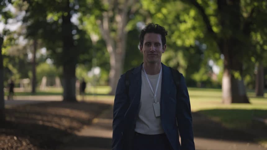A man is walking through a park on a sidewalk lined with trees as people walk in the distance