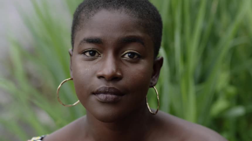Young woman with short hair and large earrings stand near a river looking at the camera