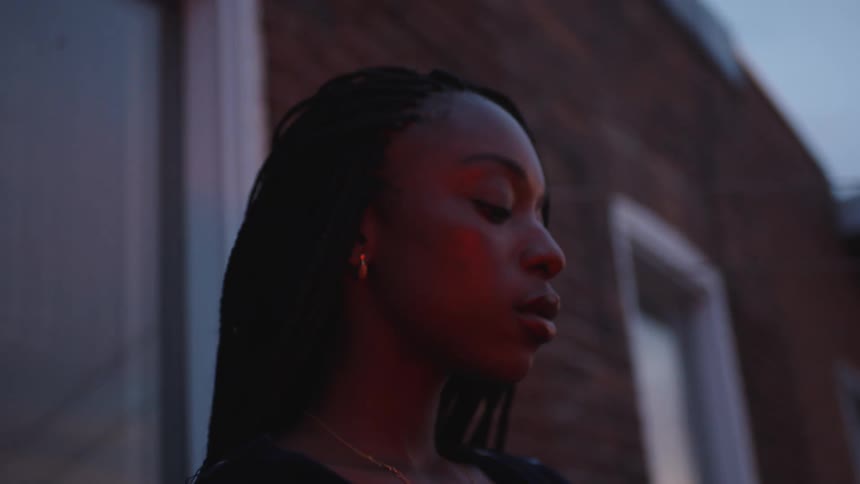 Woman with braids earrings necklace and black blouse smokes a cigarette in front of building at dusk