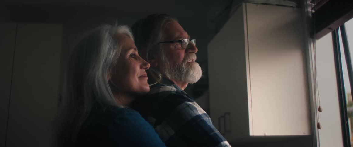 Woman back hugs ban while they look out kitchen window and talk