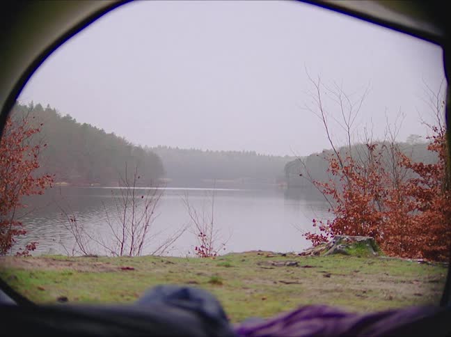 Two people in sleeping bags have the door open on their tent looking out at a lake in the mountains