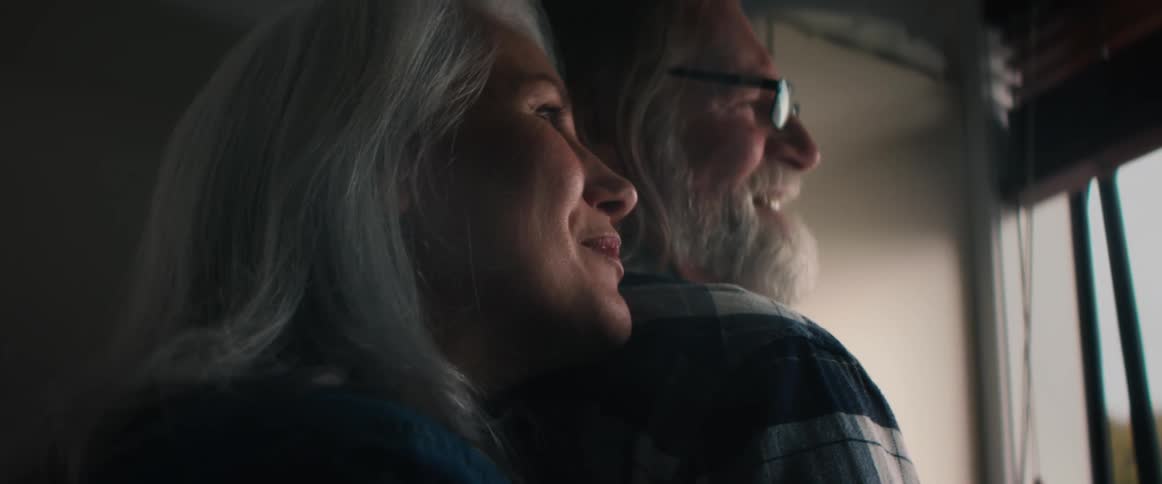 A woman hugs her husband from behind and rest her head on his back as they talk and laugh while looking out the window