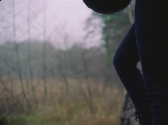 Woman with dark jeans wears black motorcycle helmet standing next to tree in the field