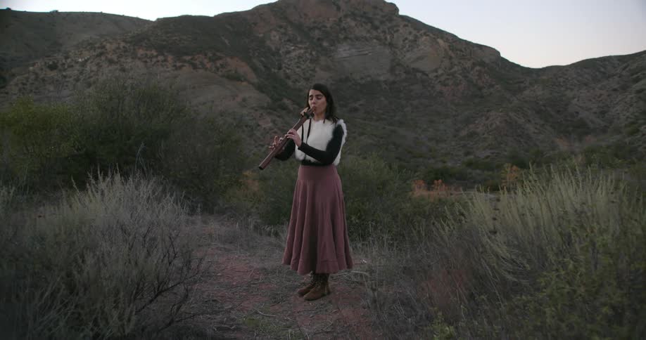 A woman wearing a long skirt is stanidng outside near a mountain playing a clarinet